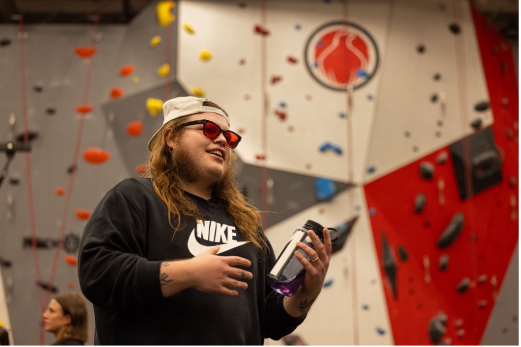 Marcus King smiling in front of a red and yellow rock wall wearing a black nike hoodie and baseball cap