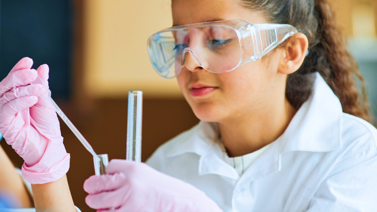 A woman with curly brown hair wearing a lab coat and goggles and pink plastic gloves holding a dropper and vial