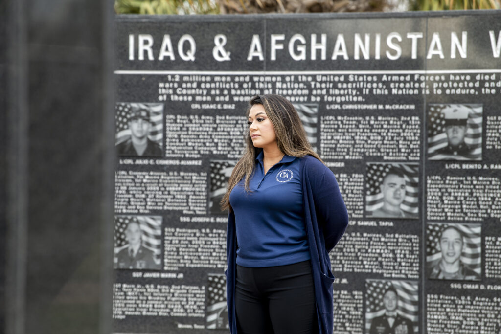 Woman in blue attire standing contemplatively beside a memorial wall dedicated to Iraq & Afghanistan veterans.