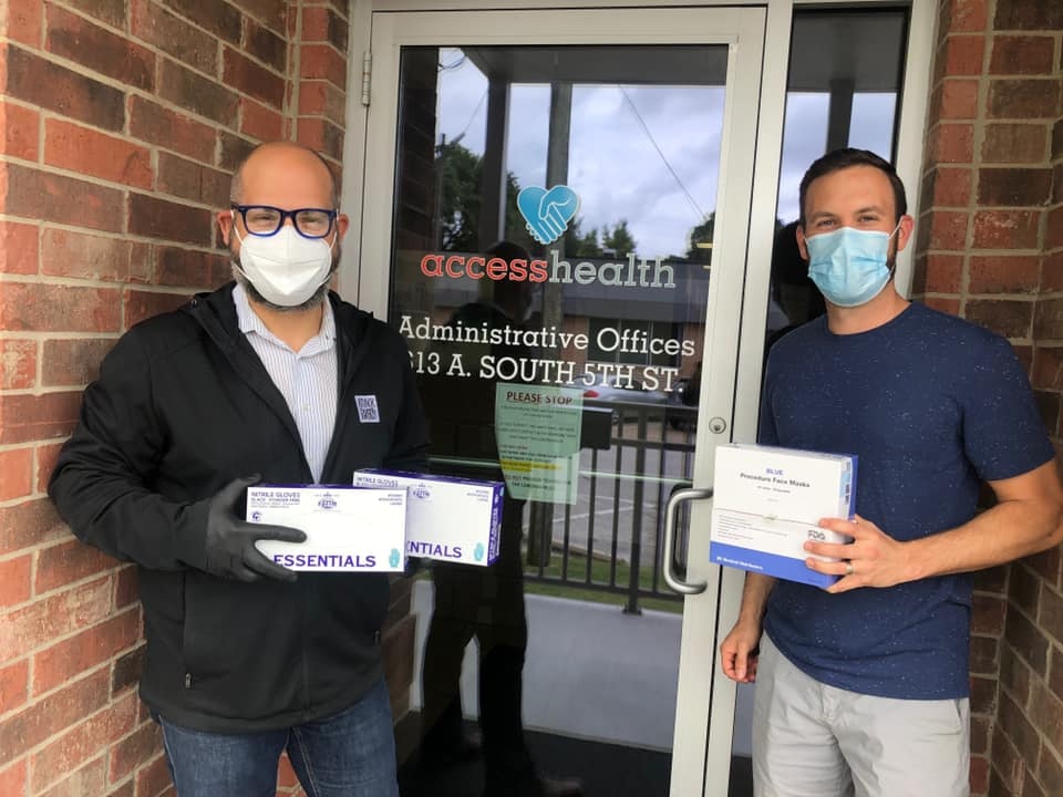 Two men with masks pose in front of an access health building with boxes in their hands
