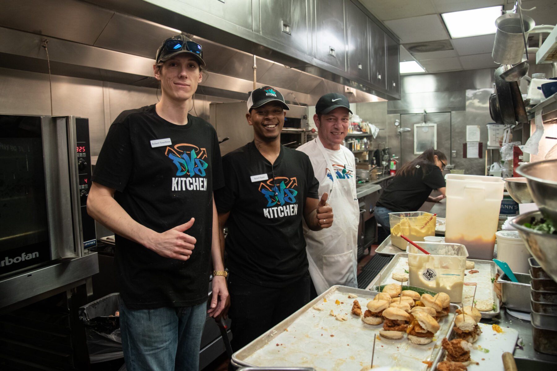 Three chefs smiling in a busy kitchen, wearing 'DV8 Kitchen' shirts.