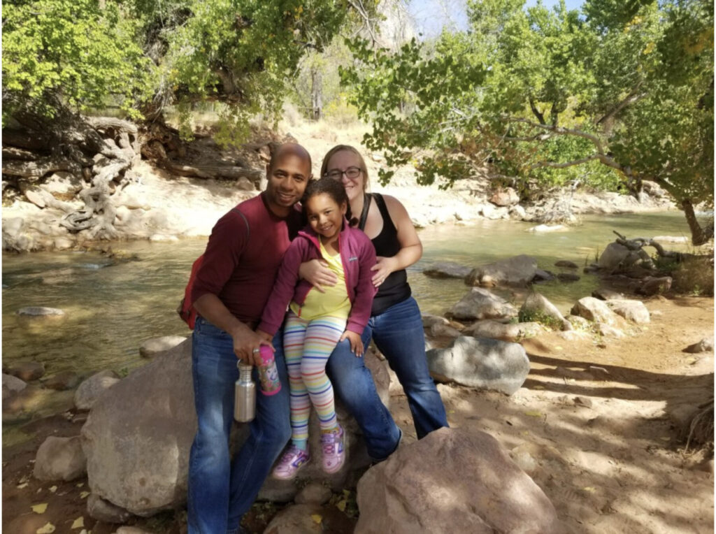 A family of three posing by a stream, surrounded by trees and boulders, smiling at the camera.