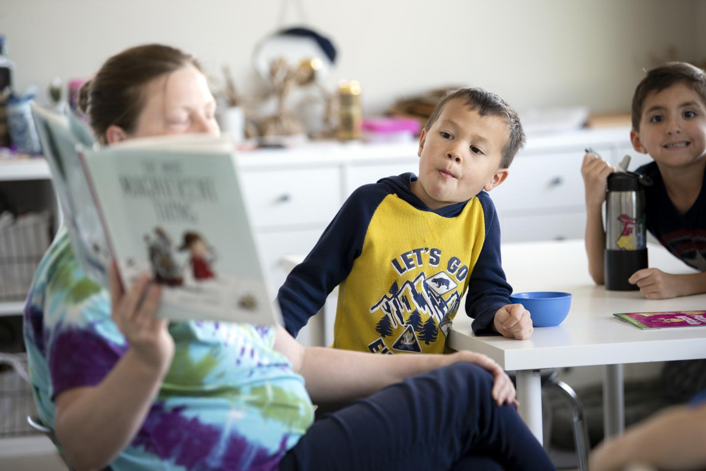 Two young boys attentively listen as a woman reads from a book titled 'The Night Before Kindergarten' in a classroom setting.