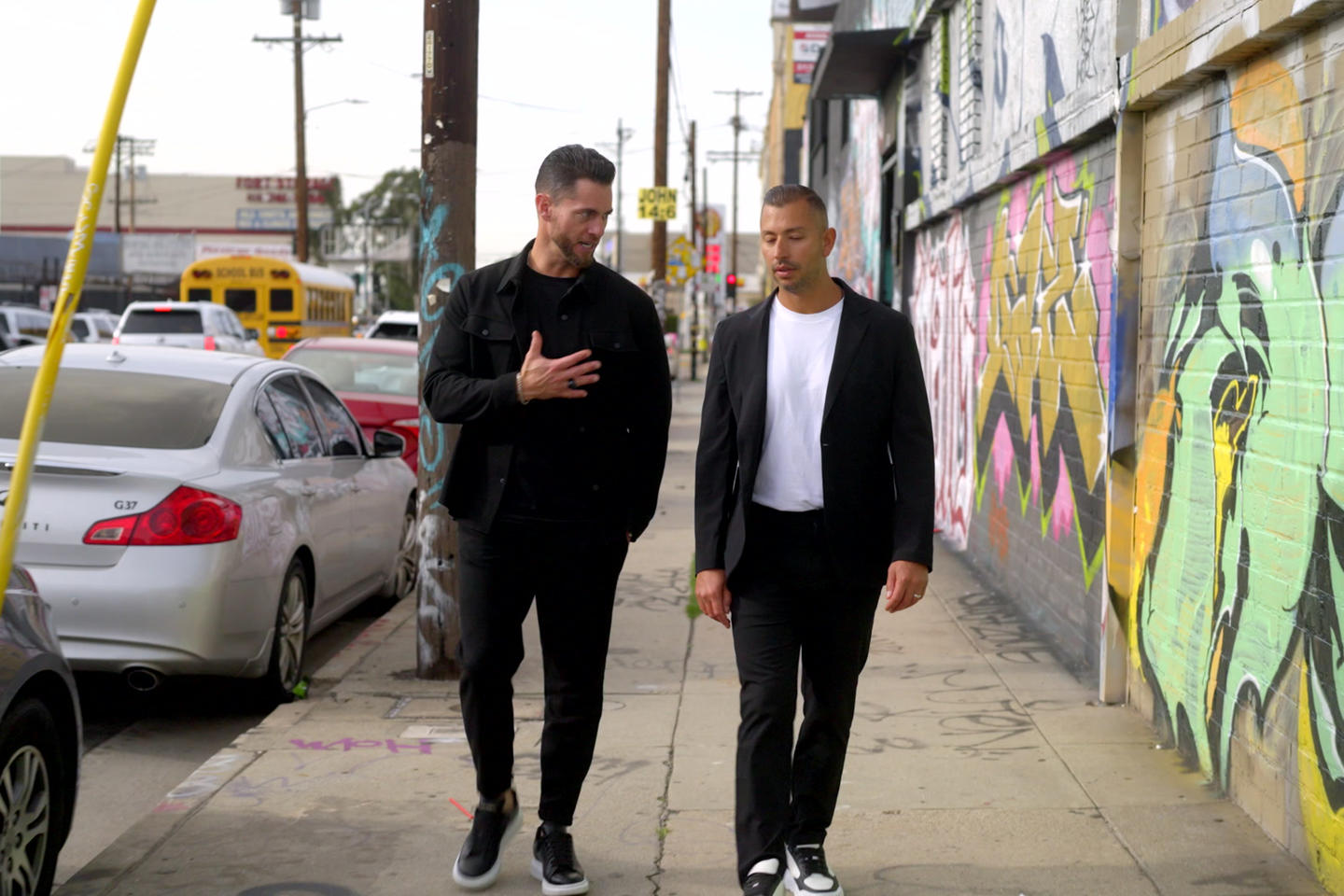 Two men walking and conversing on a graffiti-lined street with parked cars and a school bus in the background.