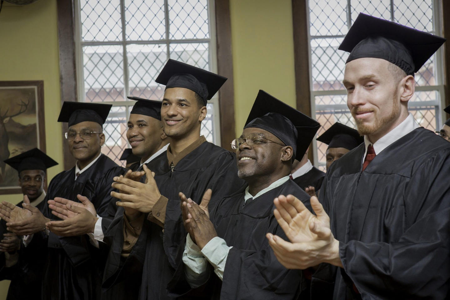 Group of smiling graduates in black robes and caps, clapping in a room with stained glass windows.