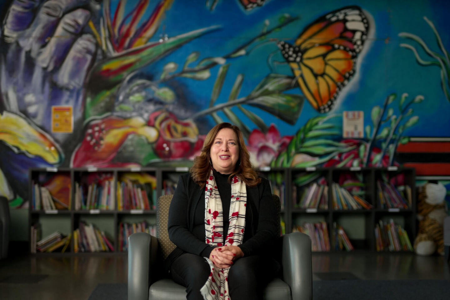 Woman sitting in front of a vibrant mural, with bookshelves and stuffed toys nearby.