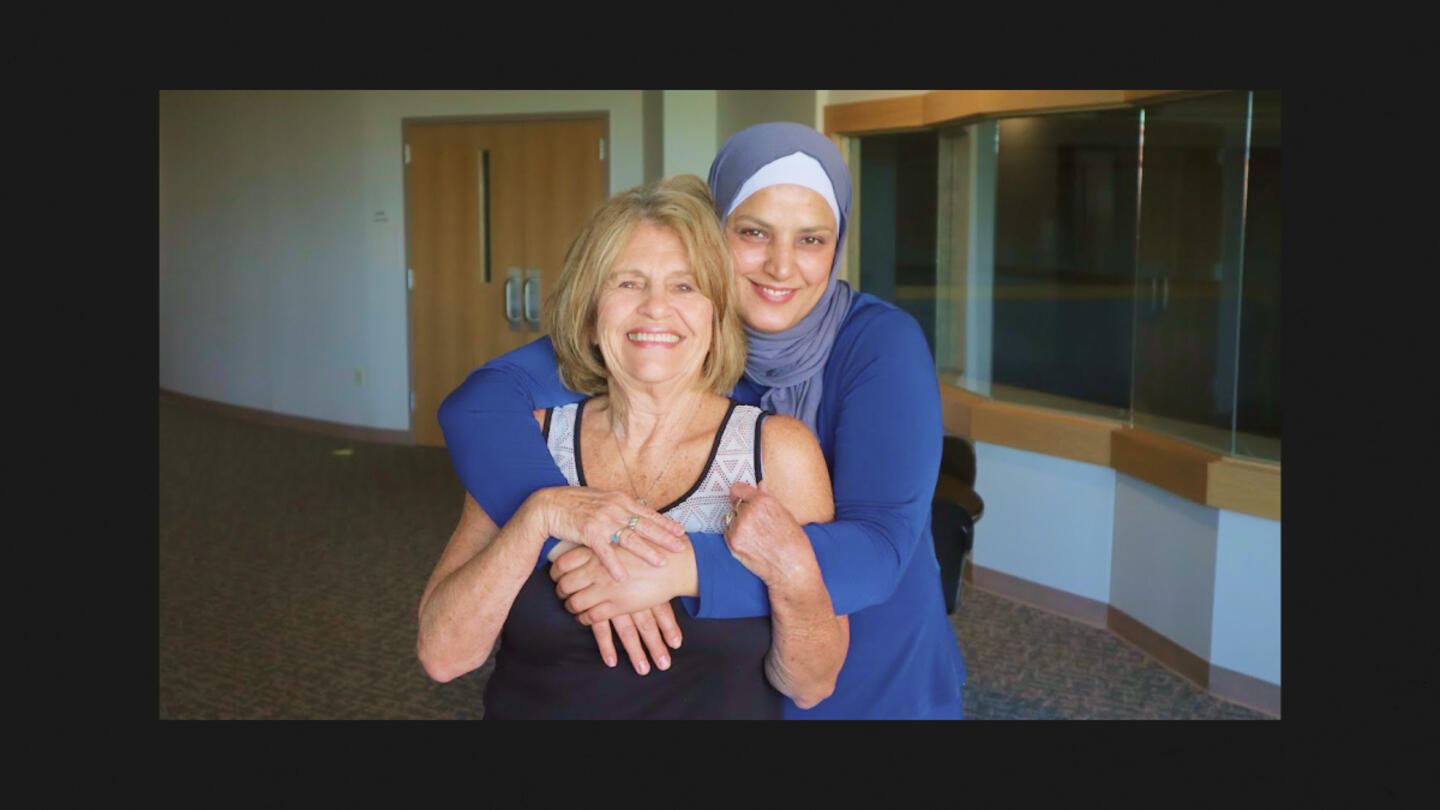 A young woman wearing a blue dress and a hijab hugging an older woman with blonde hair