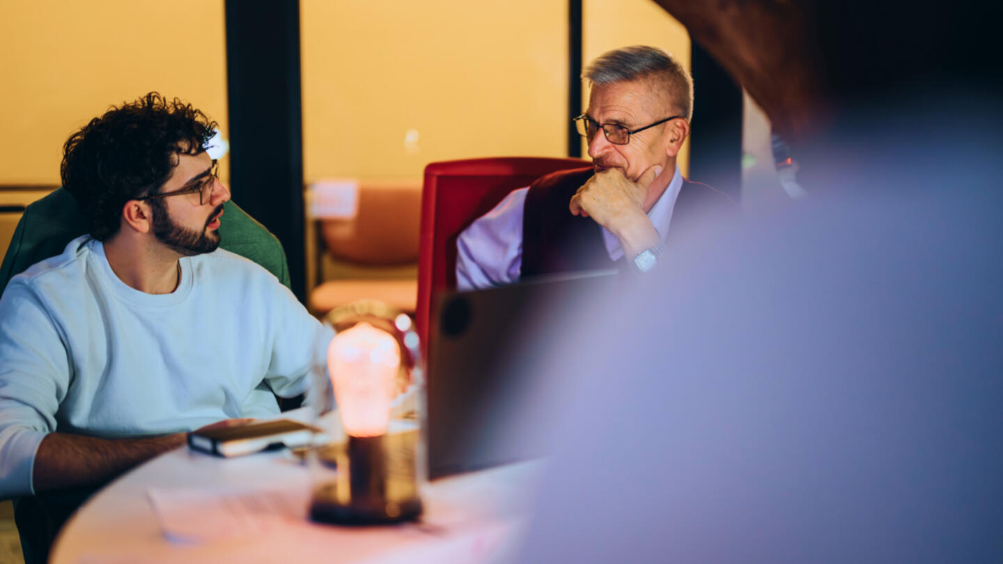 A young man with glasses and a beard wearing a light blue shirt talking to an older man wearing a vest with his hand on his chin and a laptop on the table.