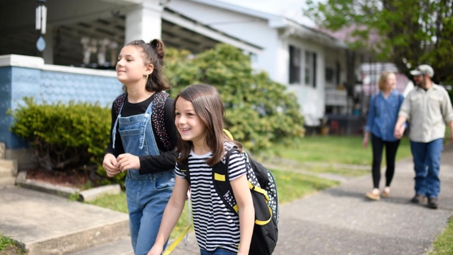 Two smiling children with backpacks walk down a sunny sidewalk.