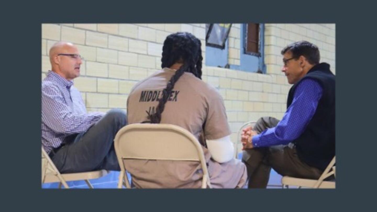 Three men sit in a discussion circle inside a jail, with one wearing a "Middlesex Jail" uniform, representing a jail rehabilitation program focused on dialogue and connection.