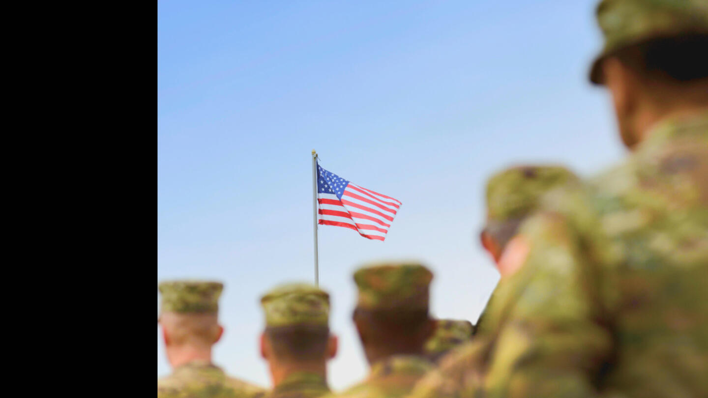 A photo of uniformed members of the military looking at an American flagshot from behind against a blue sky 