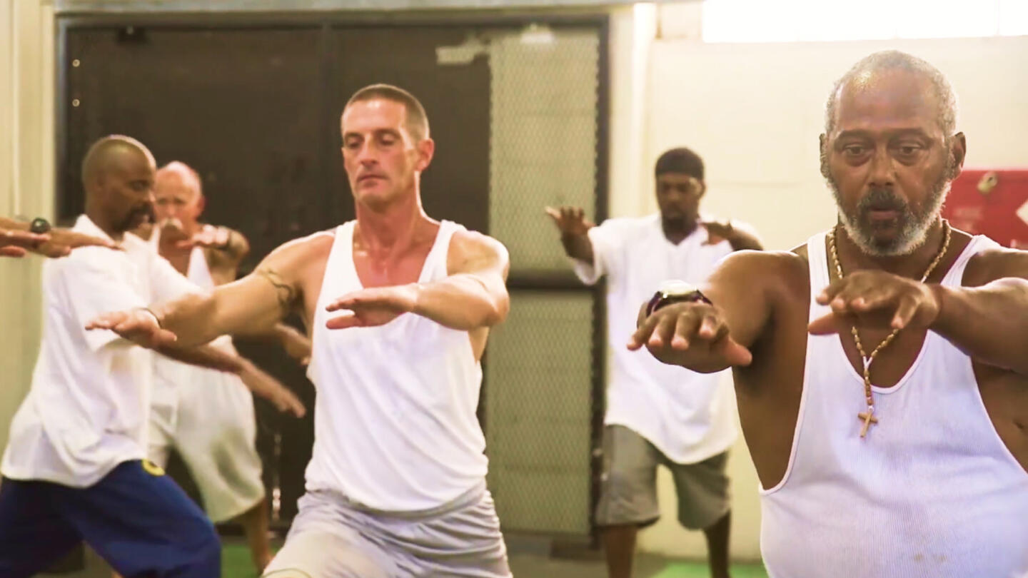 A group of men practice trauma-sensitive yoga in a gym-like setting, standing in a warrior pose with arms extended forward. Two men in the foreground, one with a beard and a cross necklace, and the other with short hair, appear focused and calm, while others in the background engage in the same pose.