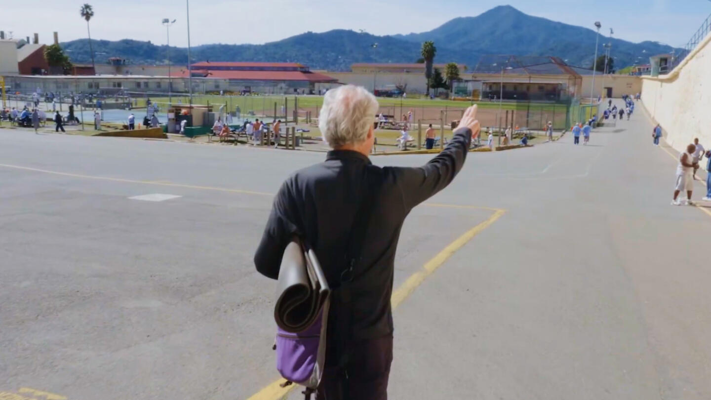 A man carrying a yoga mat points forward in a prison yard, with groups of people in the background. The setting highlights a prison yoga program under a clear blue sky.