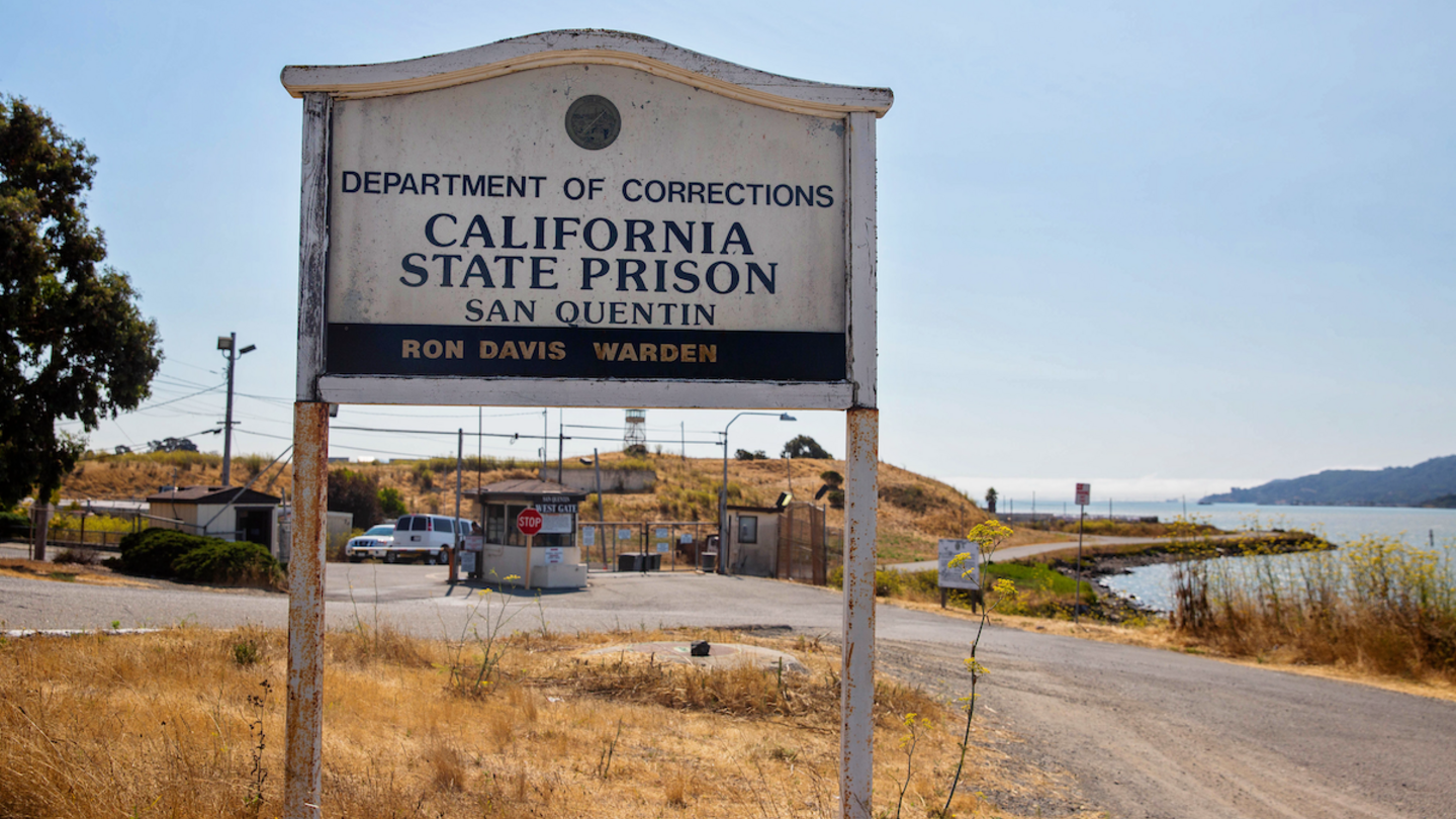 An entrance sign for California State Prison, San Quentin, sits on a dry landscape with a waterfront in the background.