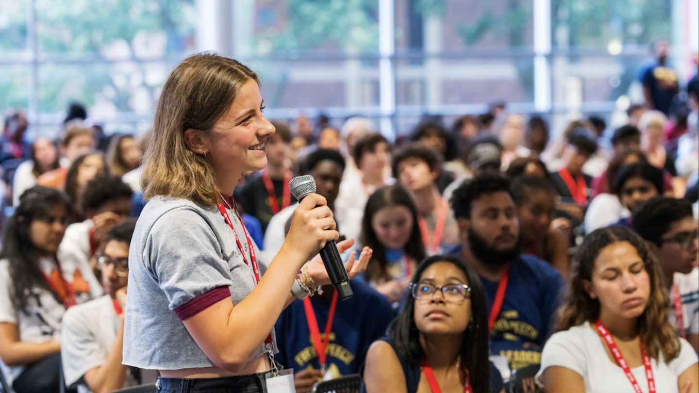 A young individual speaks into a microphone during a student event, promoting free speech and open dialogue in a diverse audience.