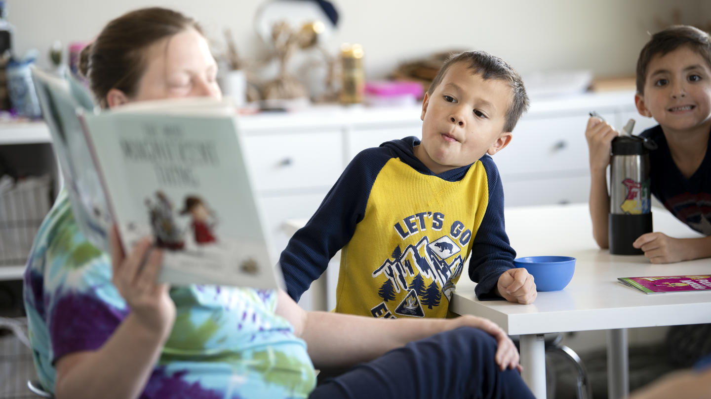 Two young boys attentively listen as a woman reads from a book titled 'The Night Before Kindergarten' in a classroom setting.