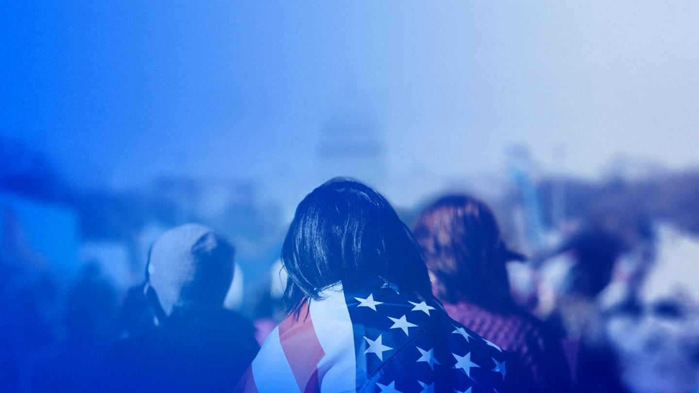A person with an American flag draped on their back like a cape amongst a crowd of people