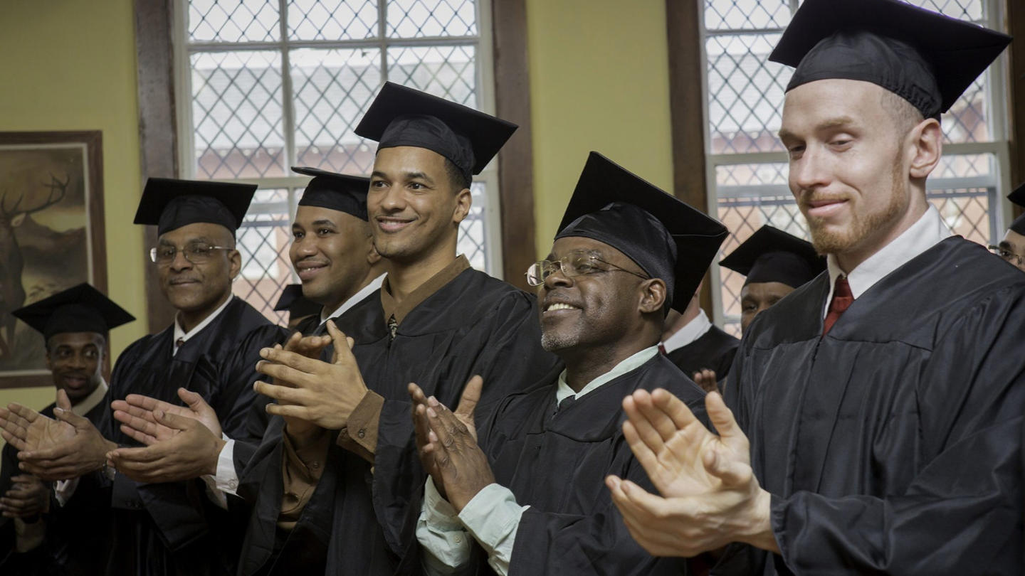 Group of smiling graduates in black robes and caps, clapping in a room with stained glass windows.