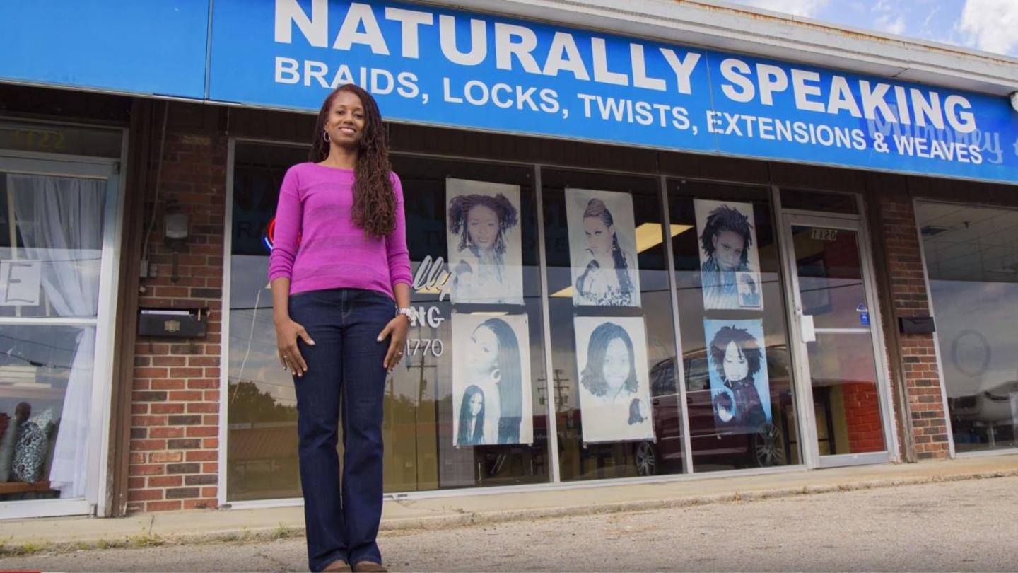 Woman in pink sweater standing in front of 'Naturally Speaking' hair salon.