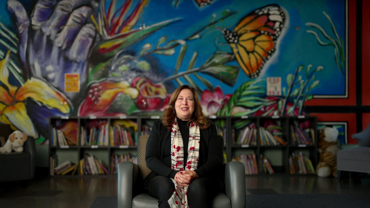 Woman sitting in front of a vibrant mural, with bookshelves and stuffed toys nearby.