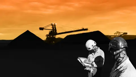 Two workers in protective gear review a clipboard in the foreground while a silhouetted mining site and machinery sit against a vivid orange sunset in the background.