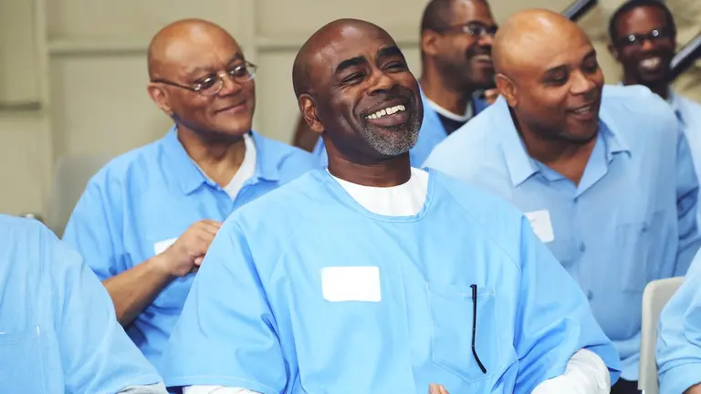 A group of incarcerated men in blue uniforms sit together in a classroom setting, smiling and engaged in conversation or a group activity.