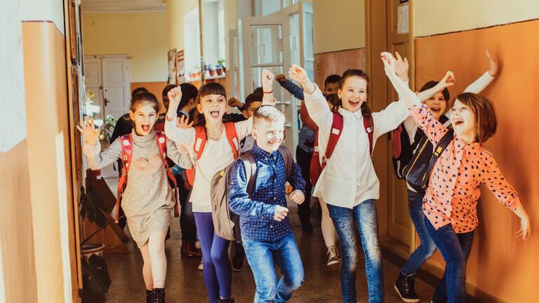 Children in a school hallway with backpacks on and their hands are in the air in excitement.