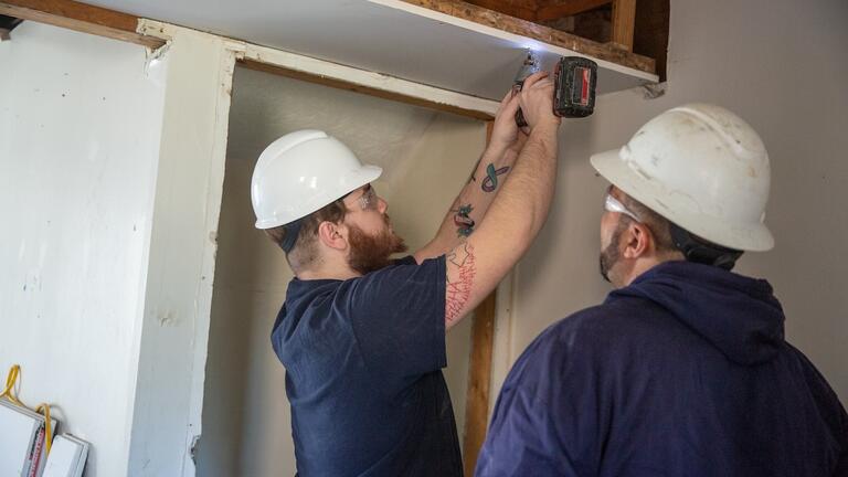 Two construction workers wearing hard hats and safety goggles install a ceiling panel, showcasing trade skills in demand that include carpentry and renovation.