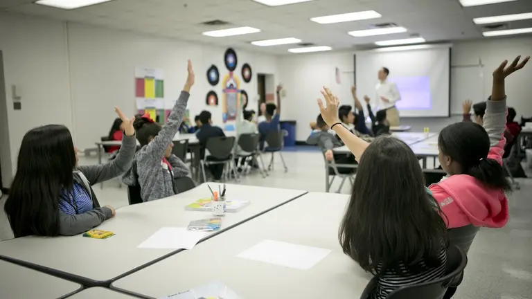 Middle school students seated in a classroom raise their hands while a teacher leads a discussion at the front.