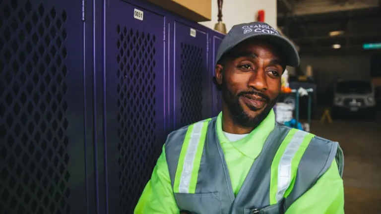 A smiling person in a reflective vest and hat stands in front of lockers.