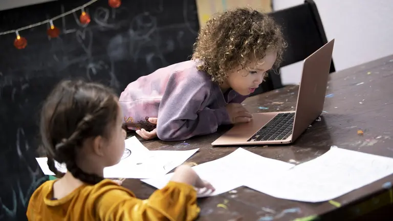 Two young children work together on school work at a table, one using a laptop while the other draws on paper.