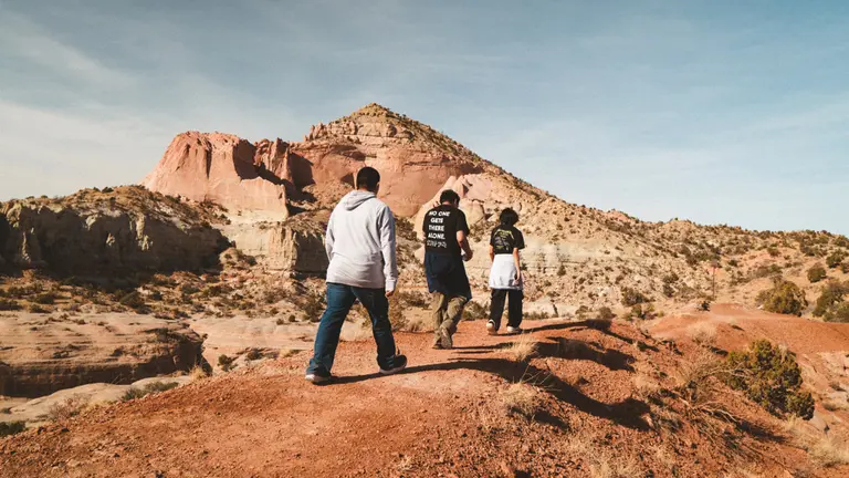 Three people walk along a red rock trail toward layered desert cliffs.