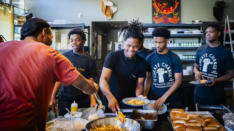 Young men work with Café Momentum, a restorative justice nonprofit,  in a kitchen, prepping and serving hot dogs and french fries.