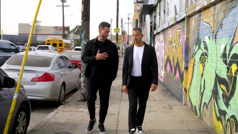 Two men walking and conversing on a graffiti-lined street with parked cars and a school bus in the background.