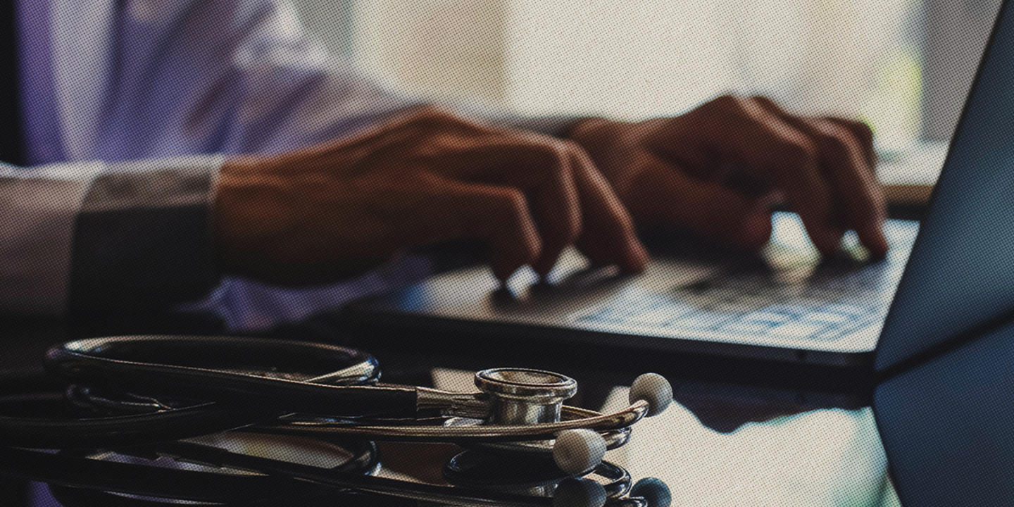 A doctor’s hands type on a laptop, with a stethoscope on the desk, representing telehealth and digital healthcare services.