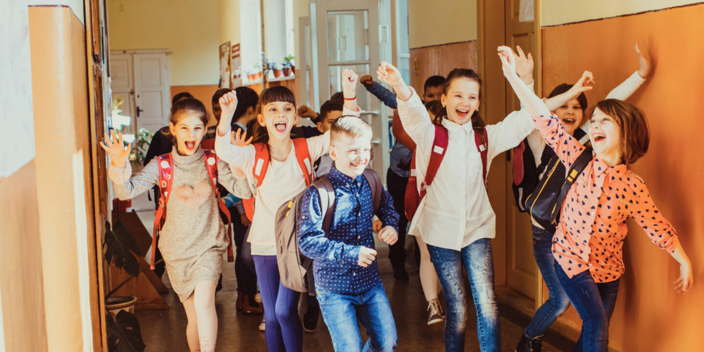 Children in a school hallway with backpacks on and their hands are in the air in excitement.