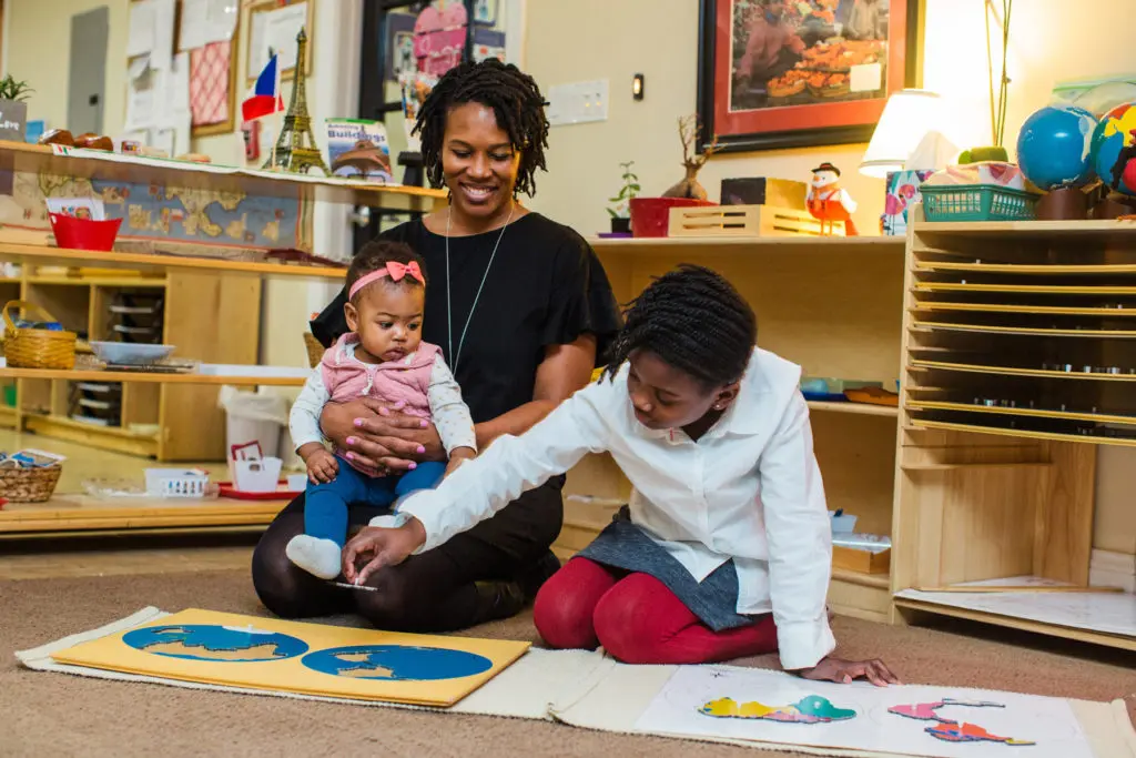Ms. Myani holds holds a baby as a young girl plays with a map game